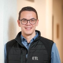 Portrait Steuerberater Torsten Warnecke mit blauen Hemd und schwarzer Weste an Wand lehnend